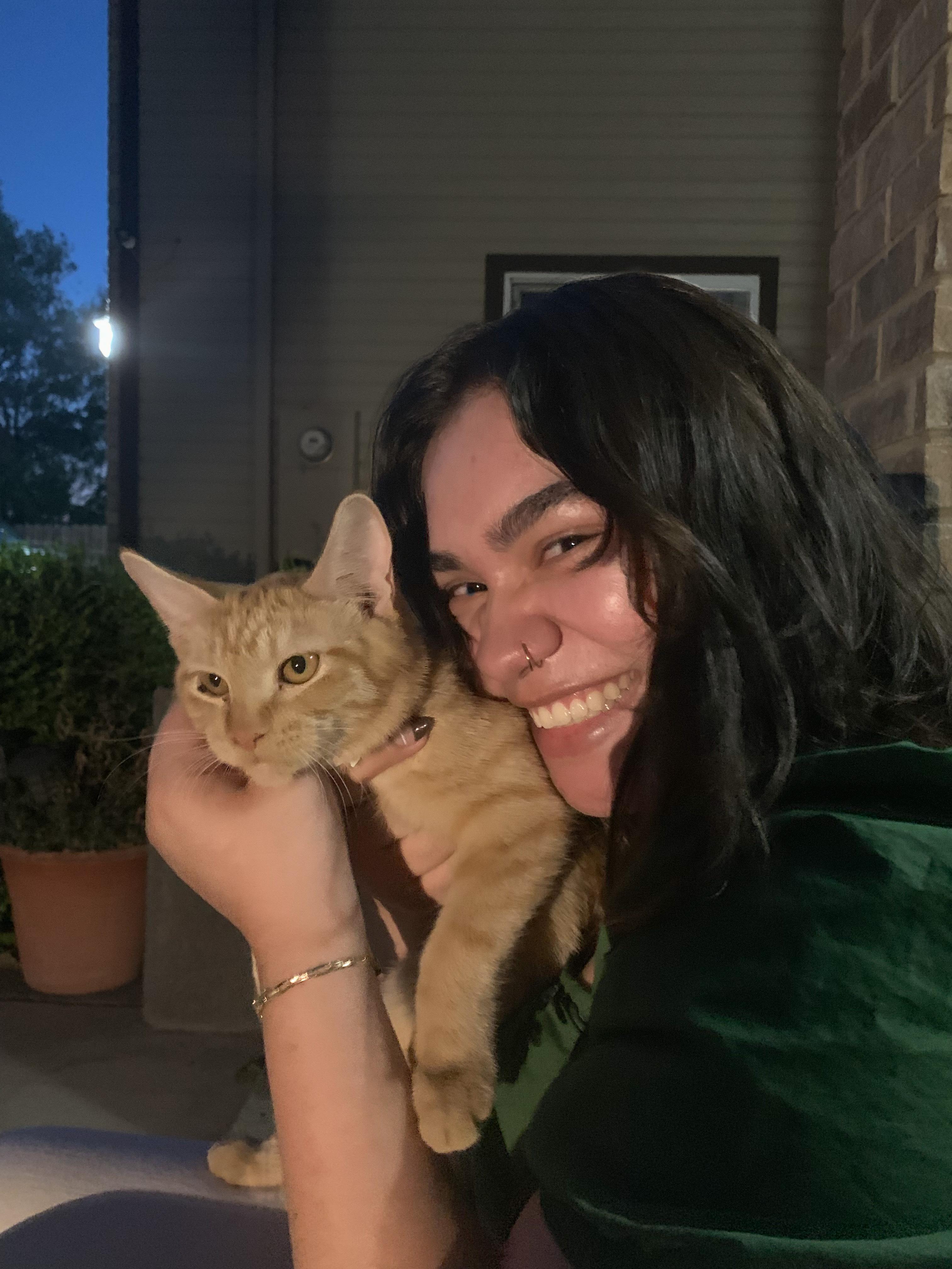 latine person with brown hair and nose ring, smiling holding orange cat, wearing green shirt and gold bracelet